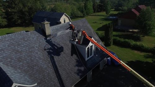 Worker using a lift to apply nanotechnology treatment on asphalt shingles, enhancing durability and lifespan, with a residential roof and surrounding greenery in the background.